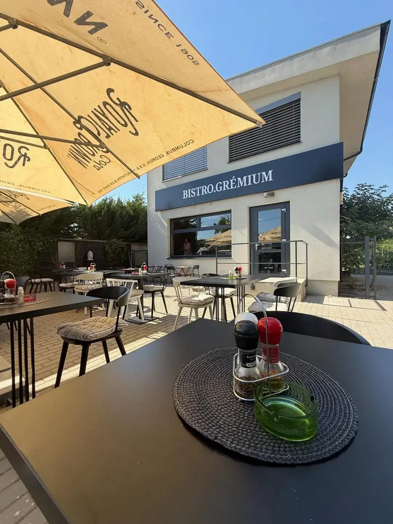 Terasa Grémia v lete, krémové slnečníky a čierne stoly pred bielym poschodovým domom s tmavomodrým nápisom Bistro.Grémium.